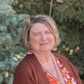 A woman with short gray hair smiling outdoors with green trees in the background.