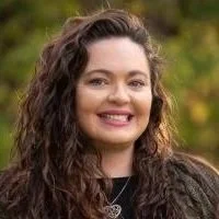 Portrait of a young woman with curly brown hair, smiling, outdoors with greenery in the background.