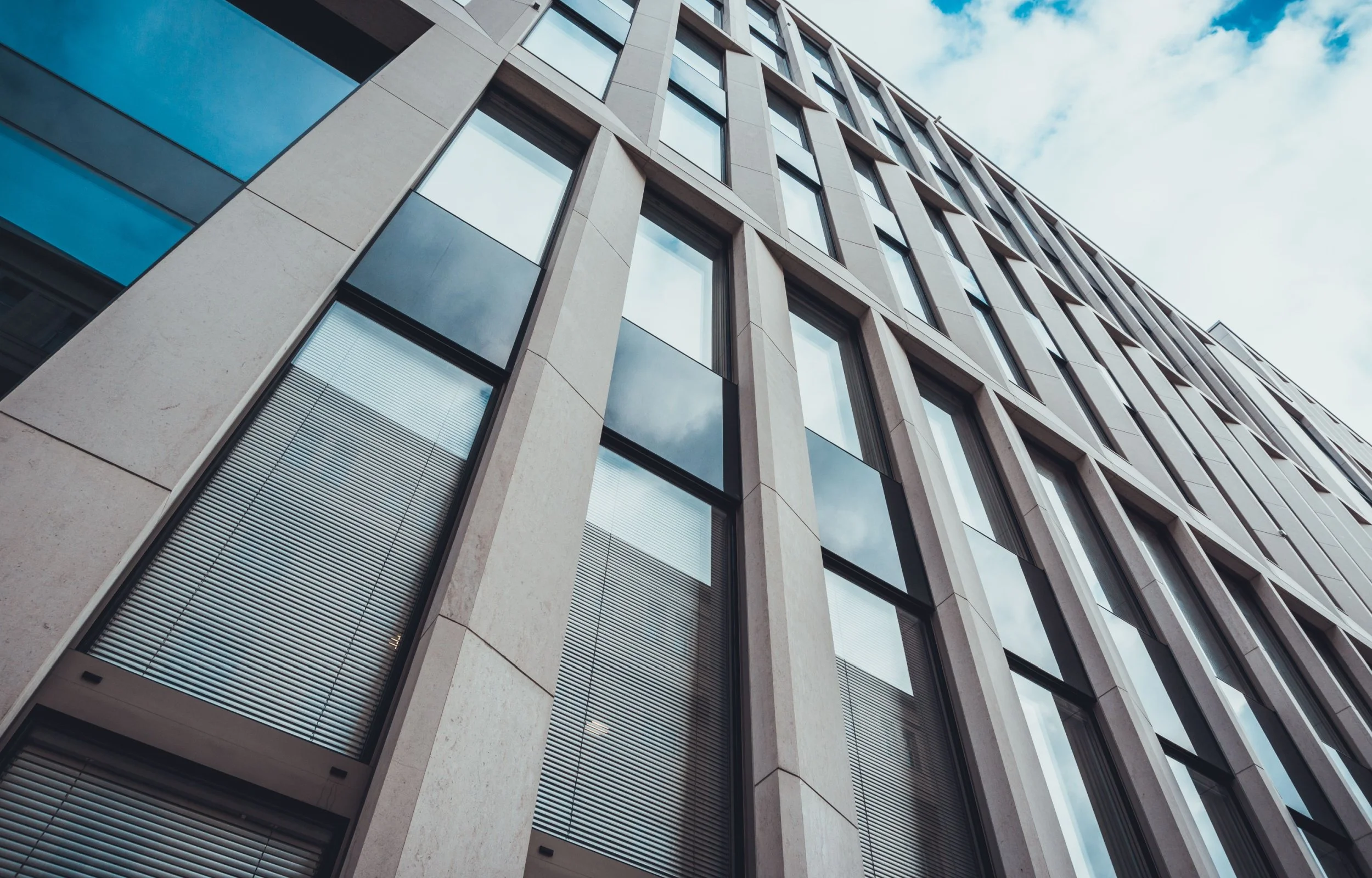 Low-angle view of a modern building with large glass windows and concrete facade, visible against a partly cloudy sky.