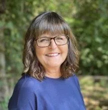 Portrait of a woman with shoulder-length brown hair, glasses, and a blue top, smiling outdoors with greenery in the background.