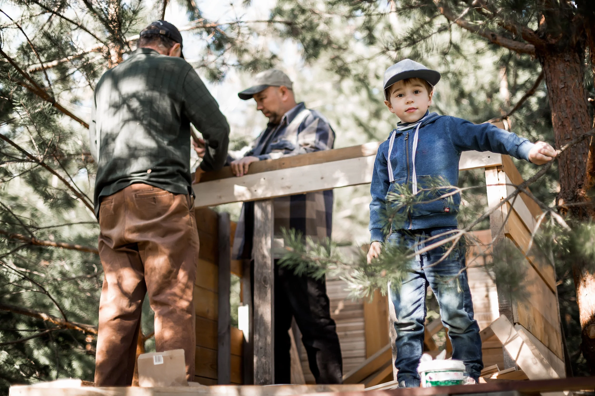 A young boy standing on a wooden platform in a wooded area during daytime, looking at the camera, with two men in the background working on a wooden structure.