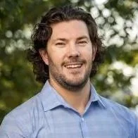 A man with dark curly hair and a beard, smiling, wearing a light blue dress shirt, outdoors with trees in the background.