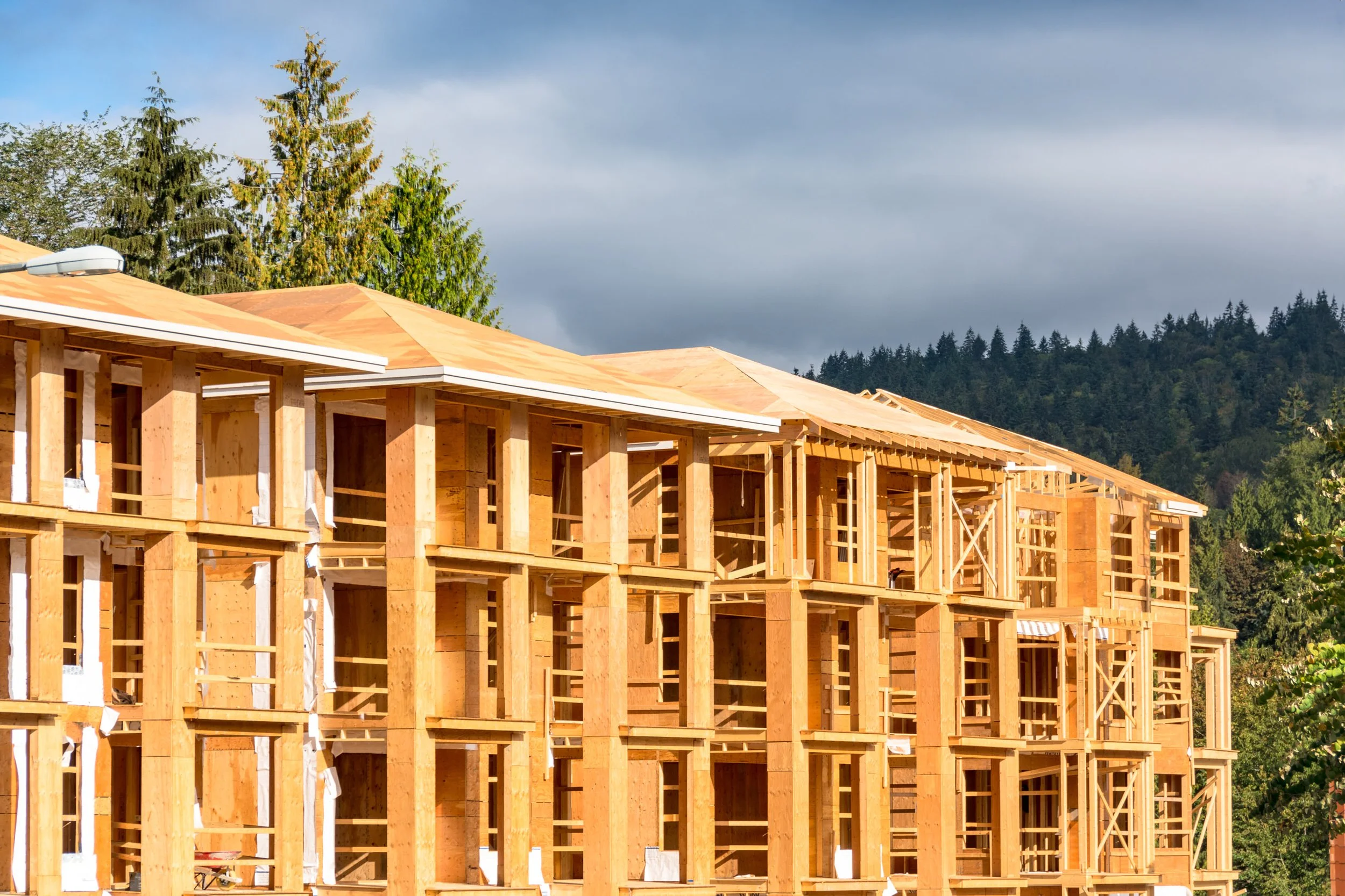 Construction site with wooden framing of residential buildings in progress, set against a backdrop of green trees and cloudy sky.