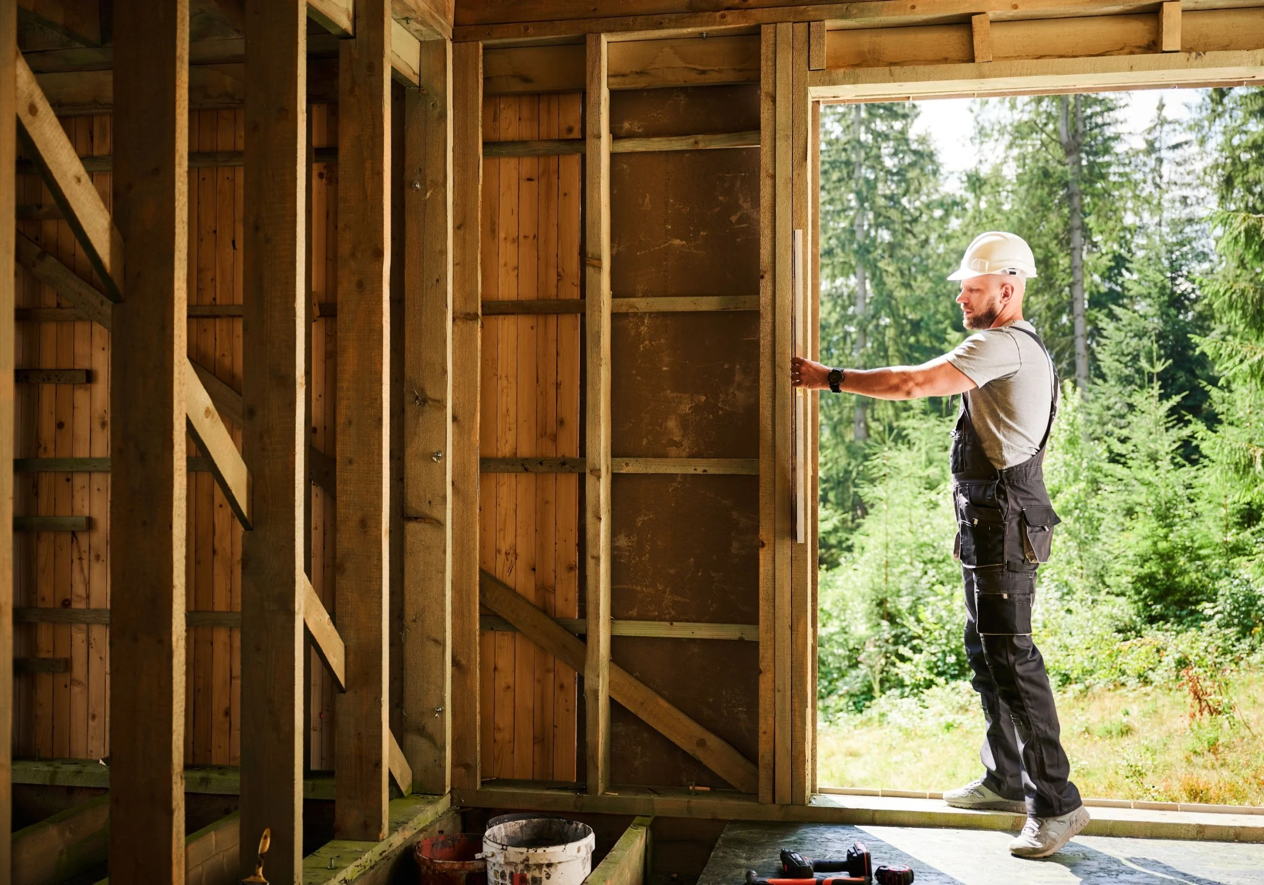 A man wearing a white construction helmet and work overalls is installing or inspecting something on the wall of a wooden structure. The overall setting is outdoor, with green trees and natural light.