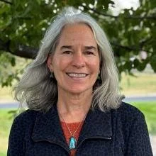 Smiling woman with gray hair outdoors, wearing a dark jacket and necklace, in front of green trees.