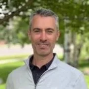 A man with short gray hair and a beard, wearing a light gray jacket and standing outdoors in a park with green trees.