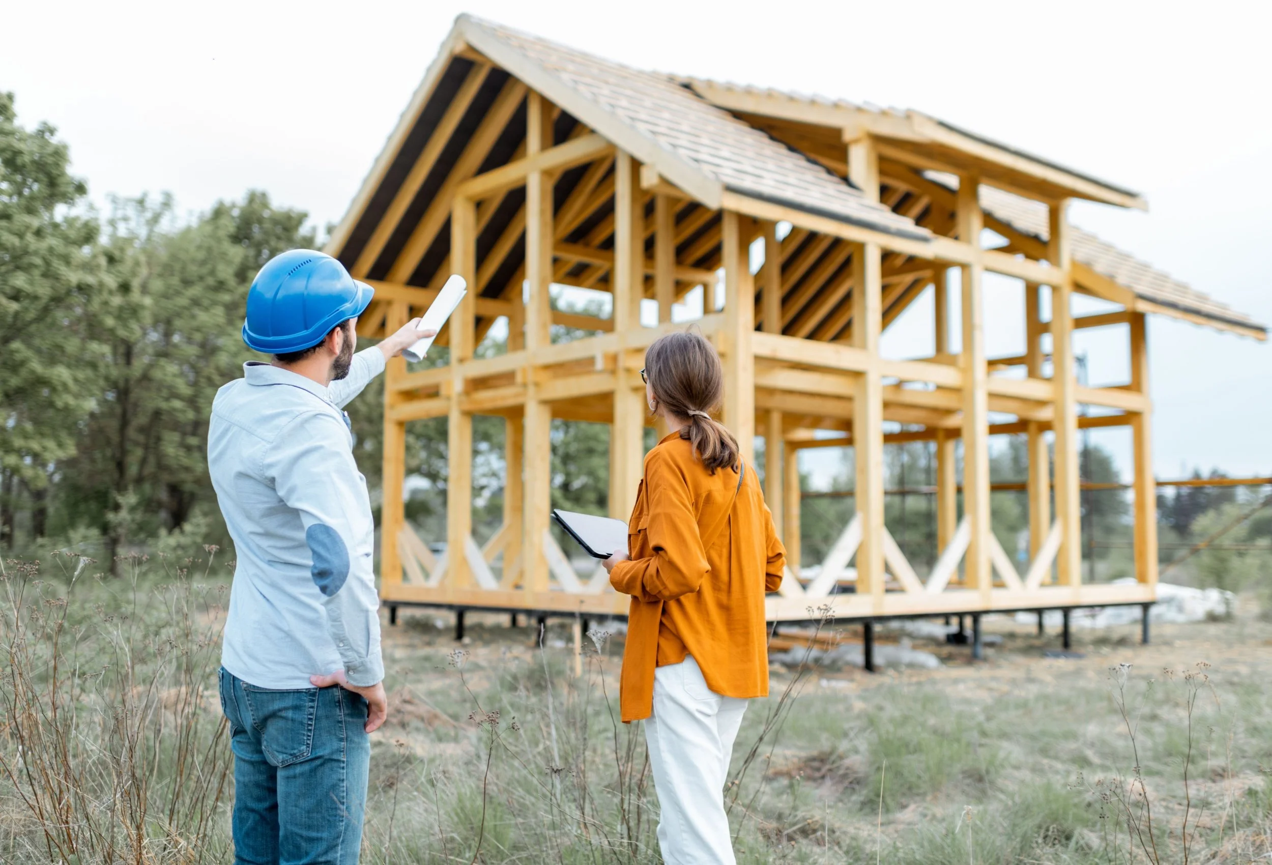 Two people, a man wearing a blue hard hat and a woman with glasses, stand in front of a wooden house under construction. The man points at the house with a white rolled-up document, and the woman holds a tablet. The house has an unfinished wooden frame and roof. Trees are visible in the background.