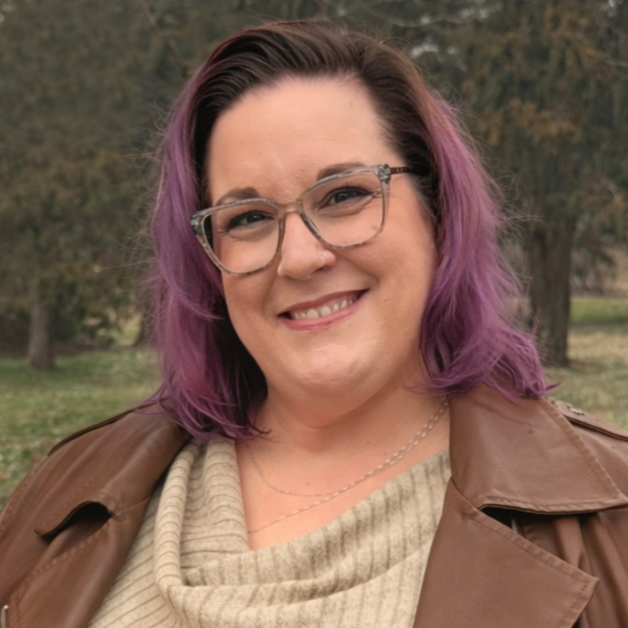 A woman with shoulder-length purple hair and glasses posing outdoors, smiling, with trees in the background.