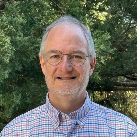 A middle-aged man with glasses and a beard, wearing a plaid shirt, standing outdoors in front of green trees.