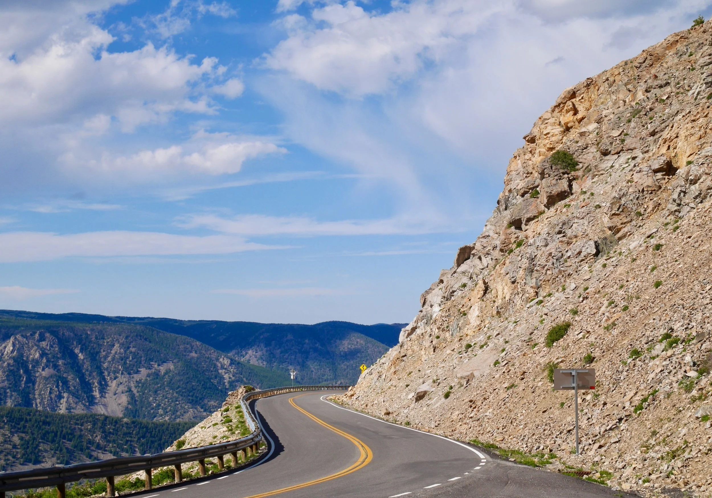 A winding mountain road with a metal guardrail on the left, cut into a rocky hillside on the right, with distant mountain ranges under a partly cloudy blue sky.