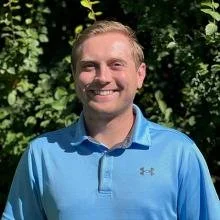 A young man with light hair smiling outdoors, wearing a blue polo shirt.