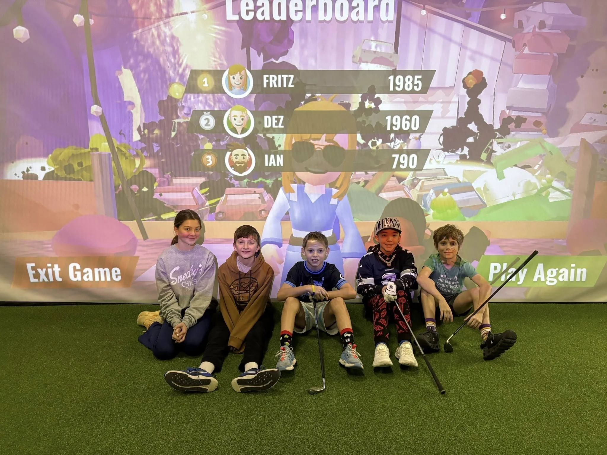 Four children sitting on the floor in front of a large screen with a leaderboard displayed. The leaderboard shows Fritz in first place with 1985 points, Dez in second with 1960, and Ian in third with 790. The children are smiling and appear to be in 