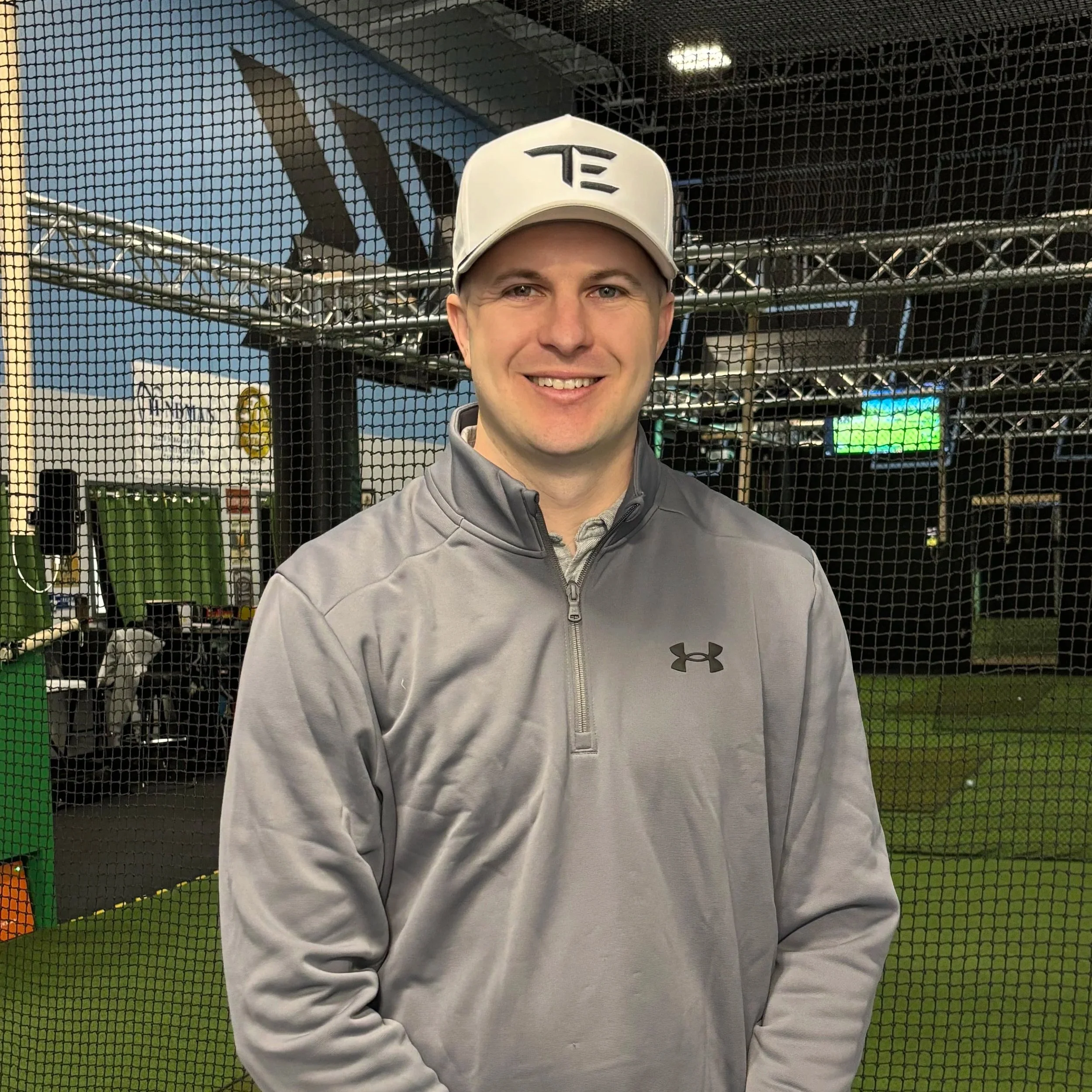 A man wearing a white cap and a gray jacket standing behind a black sports net with an indoor sports facility in the background.