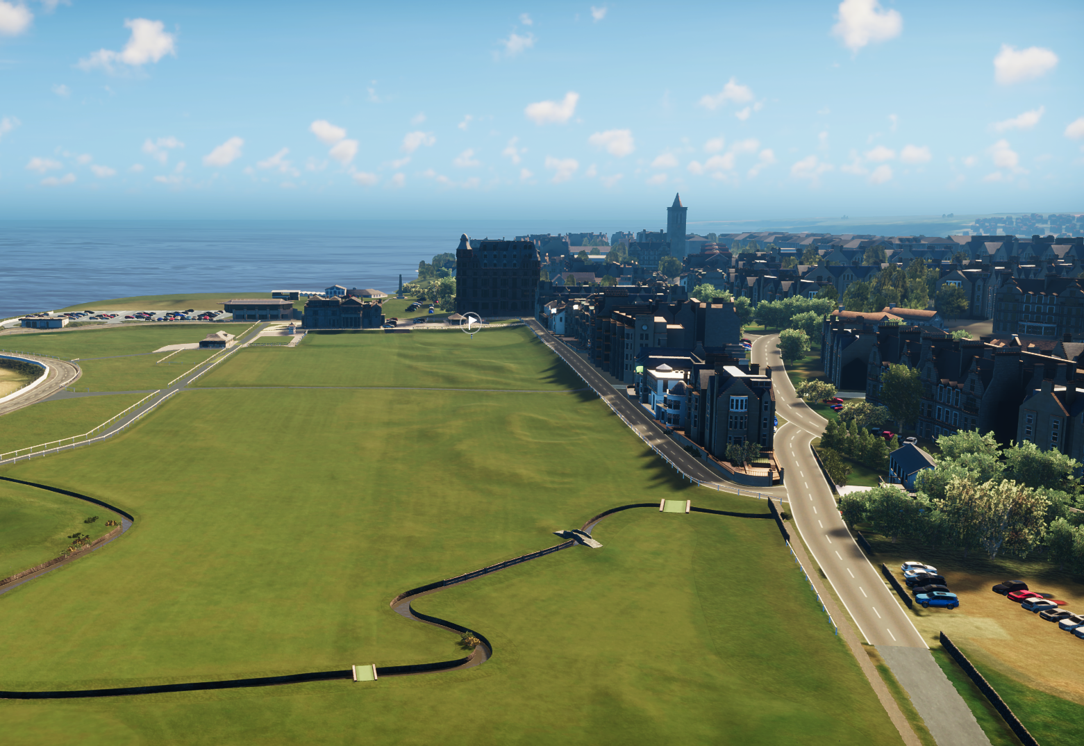 A coastal cityscape with a large grassy area in the foreground, a parking lot, and several tall buildings near the shoreline with water and clouds in the background.