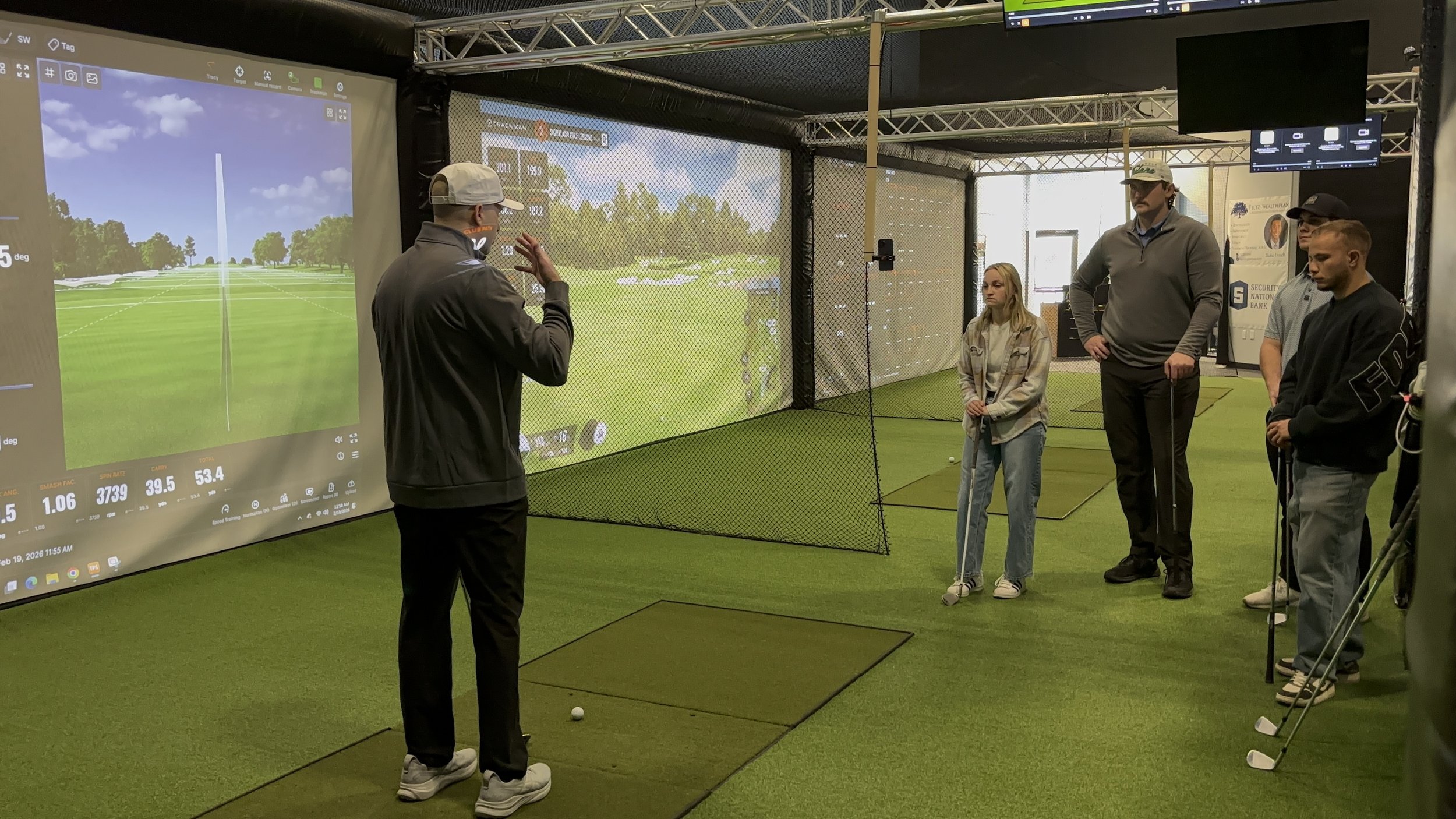 Indoor golf simulation room with a large screen showing a virtual golf course, a person in a gray jacket and white cap discussing, and four other individuals observing, some holding golf clubs.