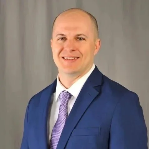 A man in a blue suit with a lavender tie, smiling, with a gray background.