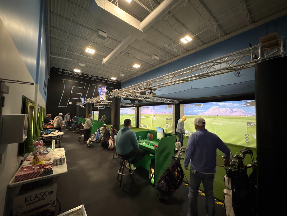 Indoor golf simulation center with multiple people playing virtual golf on large screens, with golf equipment and snacks nearby.