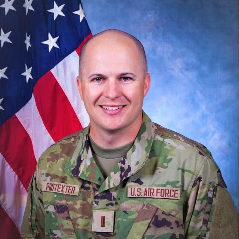 Portrait of a smiling U.S. Air Force soldier in camouflage uniform with a name tag 'Protexter' and an American flag in the background.