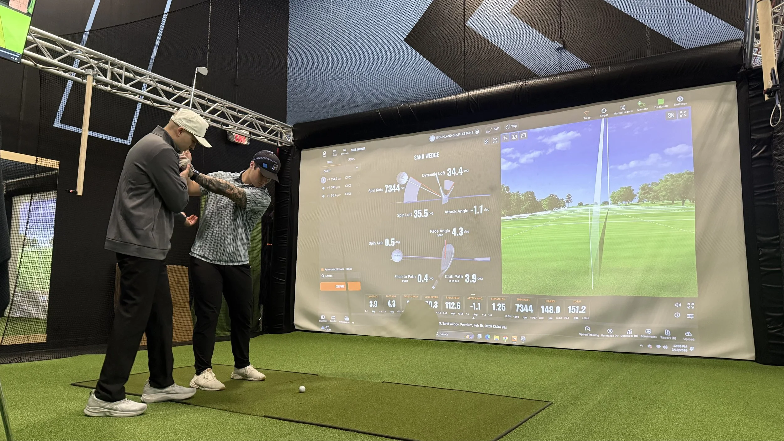 Two people practicing golf indoors using a golf simulator. One person is holding a golf club and preparing to hit a golf ball on a green artificial turf. A large screen displays golf data and a virtual golf course with a blue sky and trees.