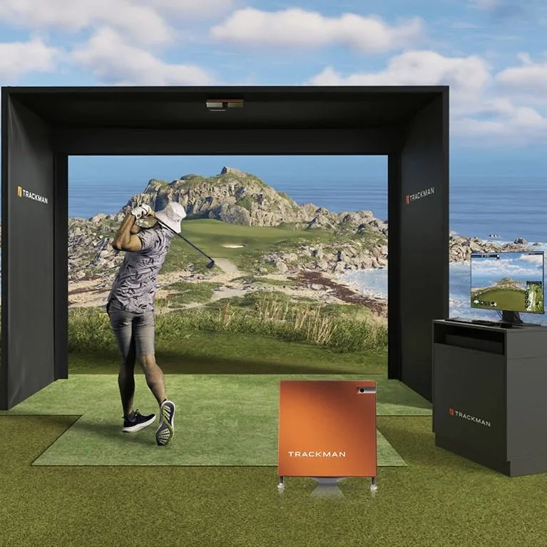 Indoor golf simulation setup with a woman swinging a golf club, a large screen displaying a golf course, and a monitor showing the same scene.