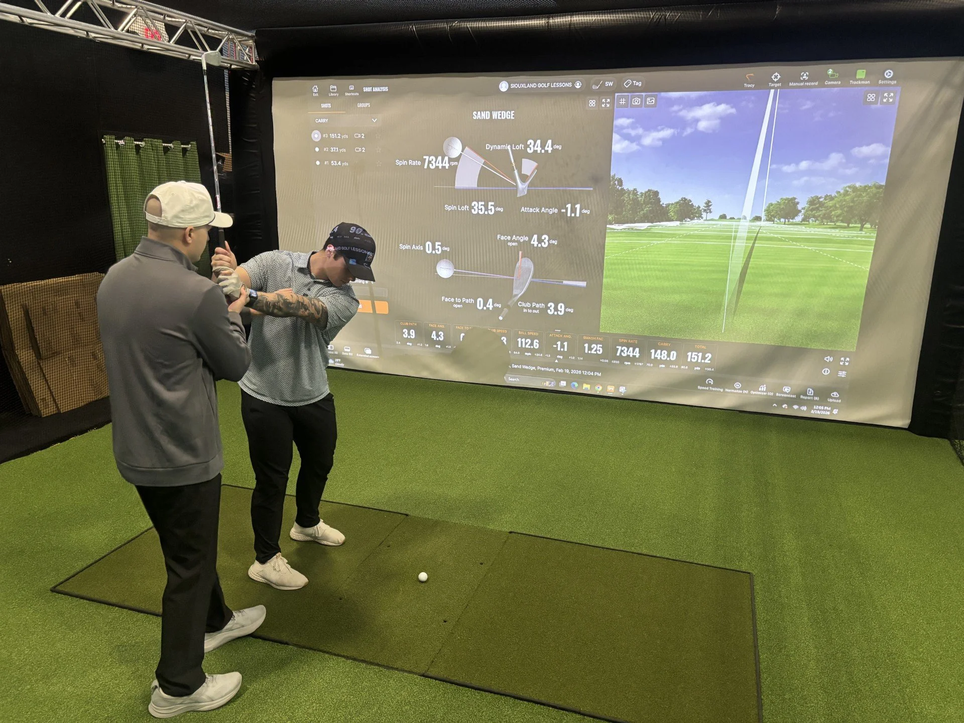 Two men are at an indoor golf simulation. One man is wearing a gray jacket and a white baseball cap; the other is wearing a black cap and a patterned shirt. They are standing on artificial green turf, looking at a large screen displaying golf swing data for a shot with a sand wedge. The screen shows details such as swing speed, launch angle, and ball trajectory.