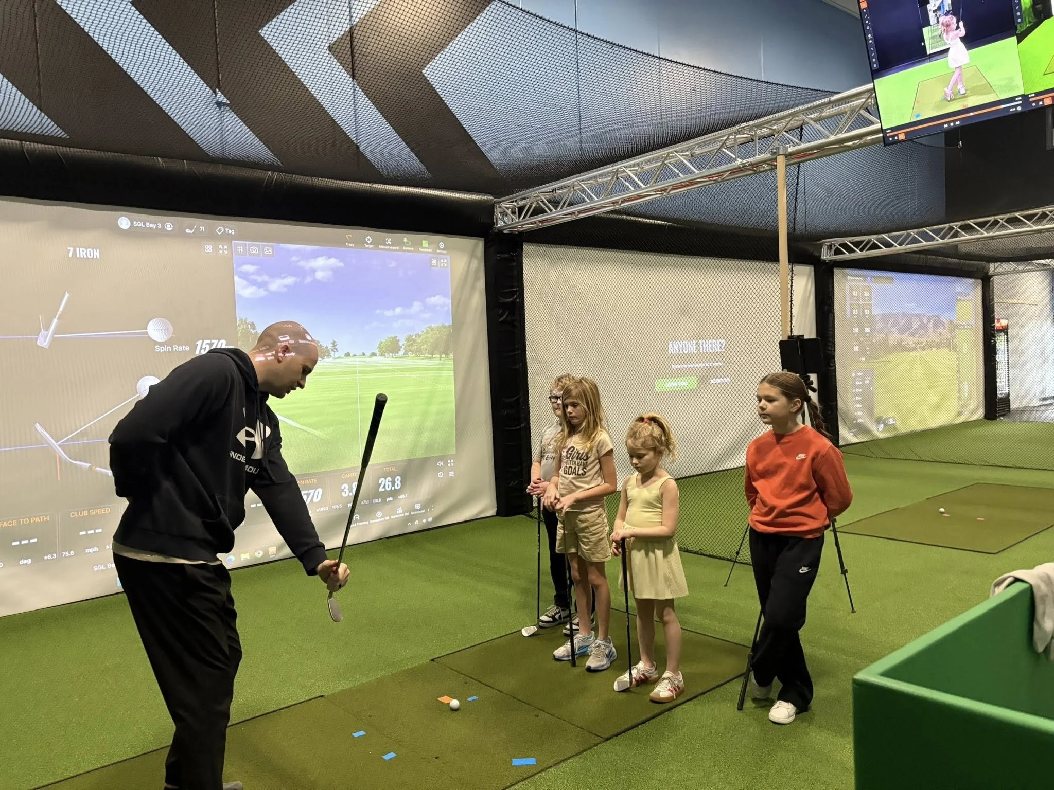 A man practicing golf indoors with a group of children observing, in front of large screens displaying a golf simulation game.