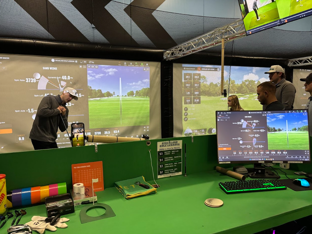 Golf simulator room with large screens displaying golf course data and a person preparing to take a shot, surrounded by a group of people watching.