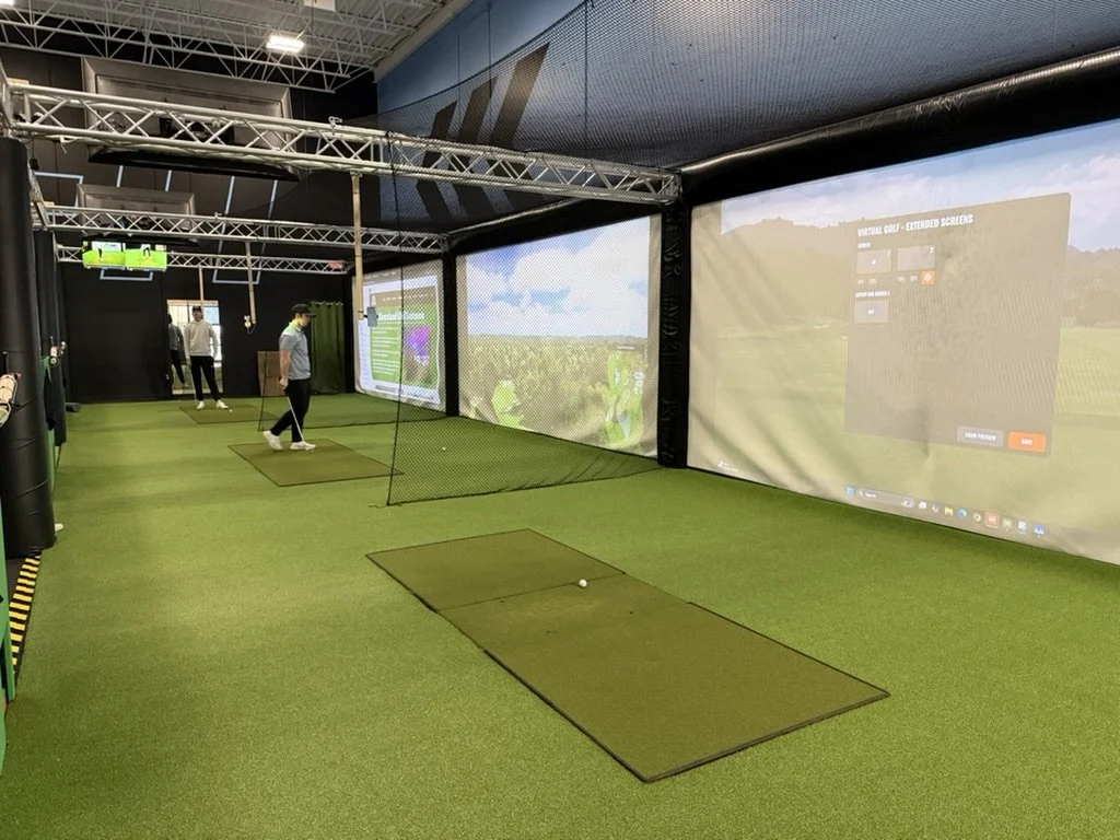 Indoor golf simulation room with artificial turf, screens displaying virtual golf courses, and people practicing golf swings.