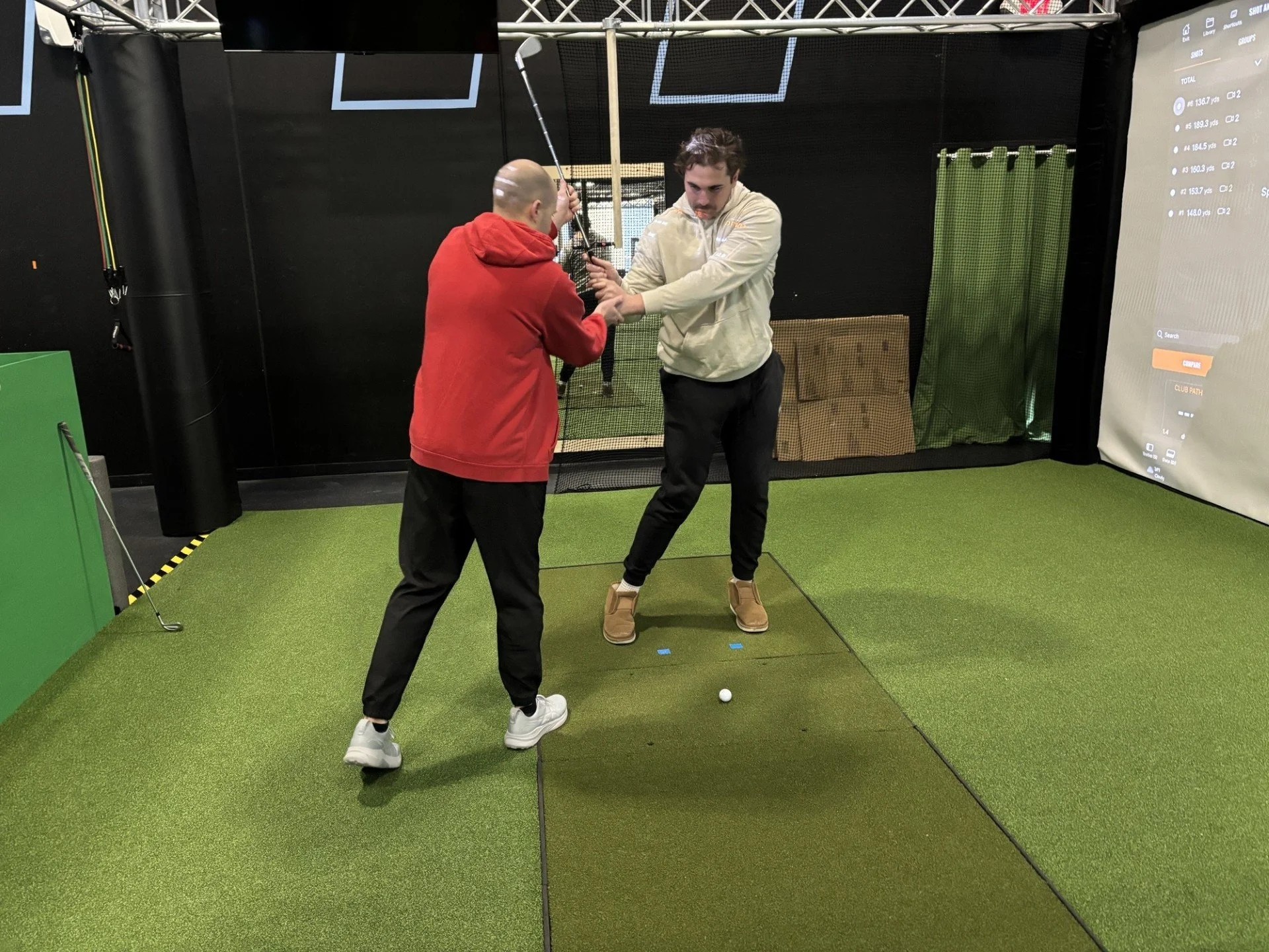 Two men playing golf indoors, one wearing a red hoodie and the other with a beige hoodie, with golf club and ball on green artificial turf.