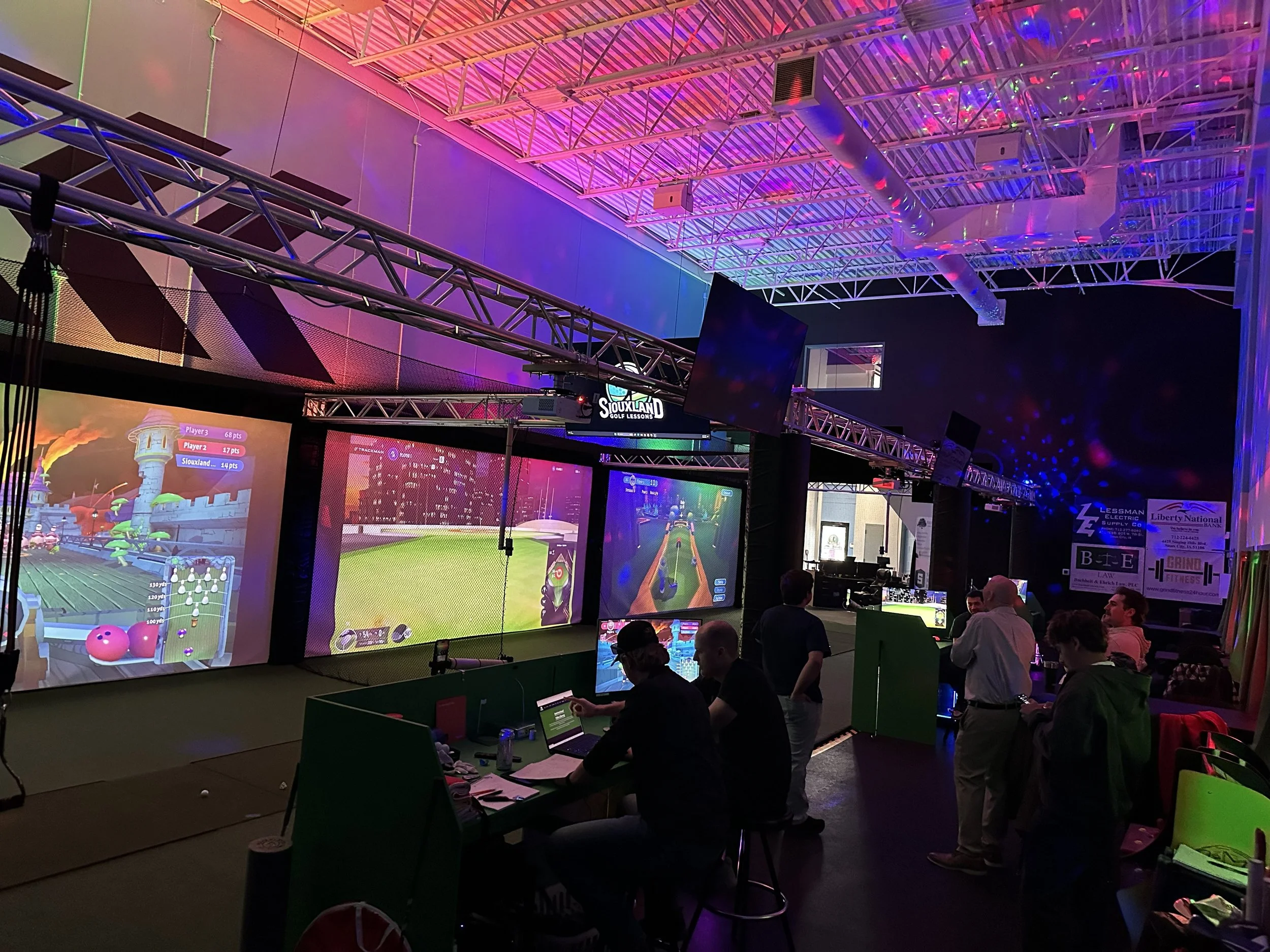 Indoor arcade or entertainment center with multiple large screens displaying digital games, colorful lights, and people playing or watching.
