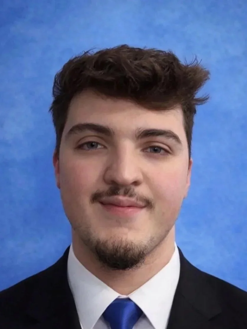 Young man with brown hair, light facial hair, wearing a white shirt, black suit, and blue tie, against a blue background.