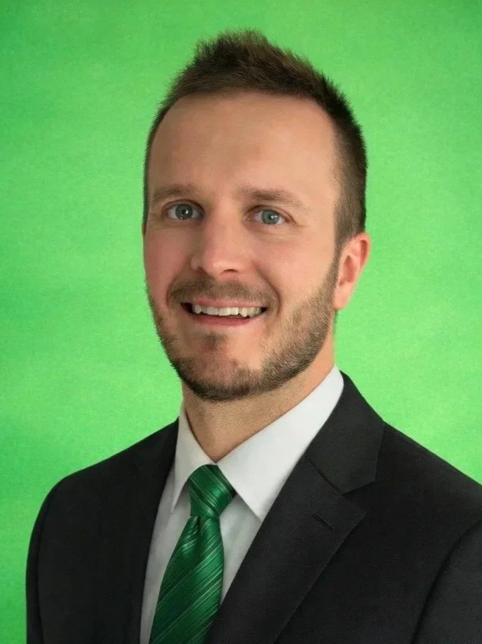 Professional portrait of a smiling man in a black suit, white shirt, and green striped tie, standing against a green background.