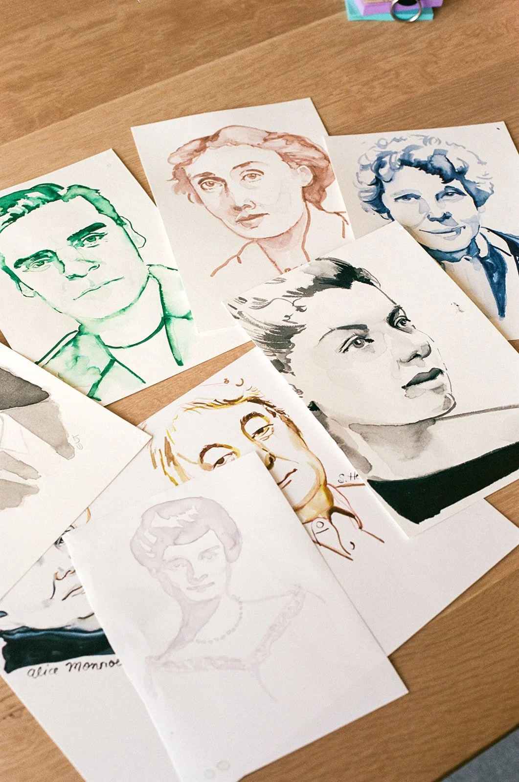 Collection of portrait sketches of various women on paper, some in color and others in black and white, spread on a wooden surface.