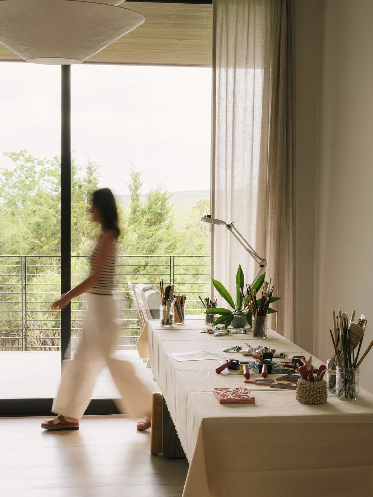 A blurred woman walking past a table set up with art supplies, including brushes, paints, and tools, on a bright room with large window and curtain, overlooking trees and a balcony.