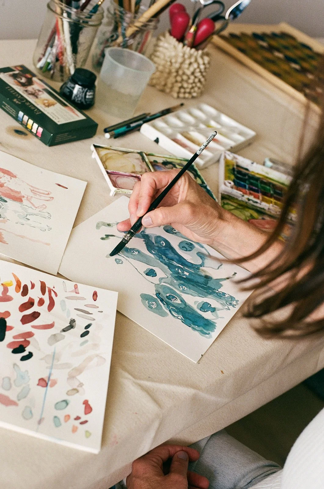 Person creating watercolor art with blues and greens, surrounded by watercolor supplies and brushes on a workshop table.