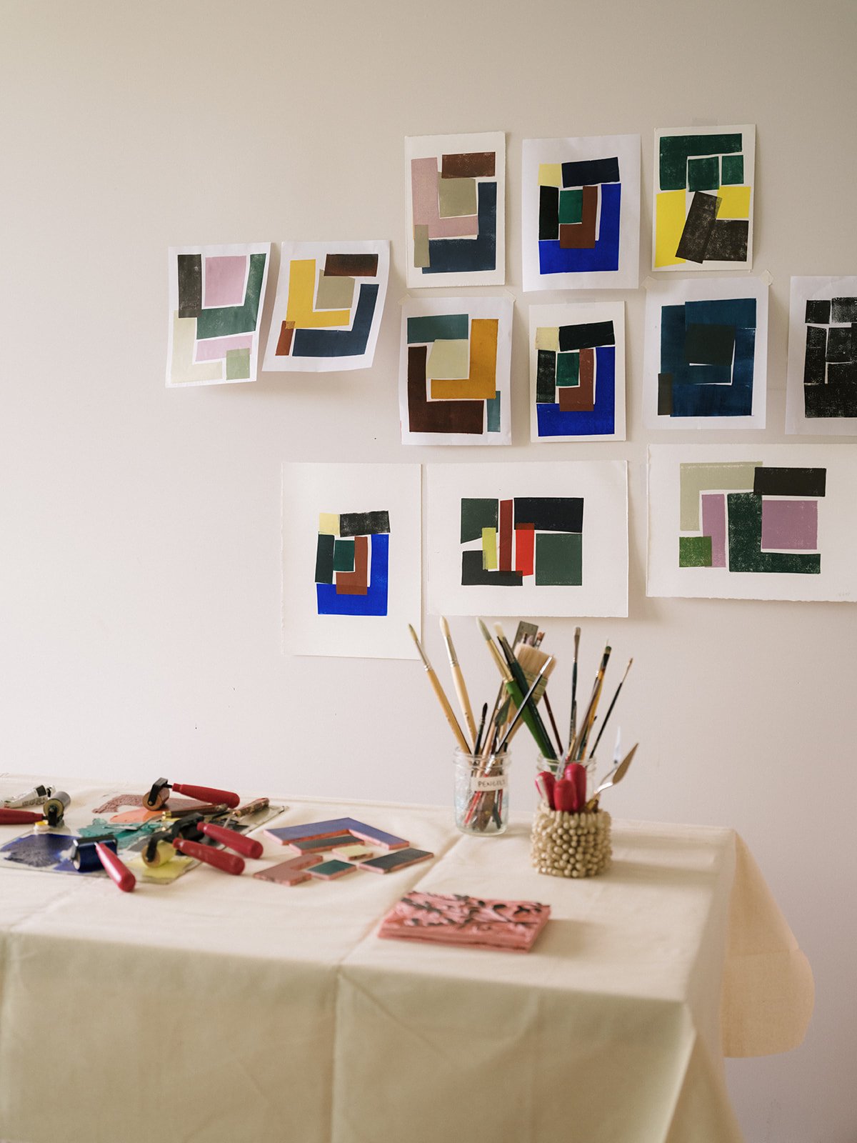 An art display with multiple colorful abstract paintings on a white wall above a table covered with art supplies.