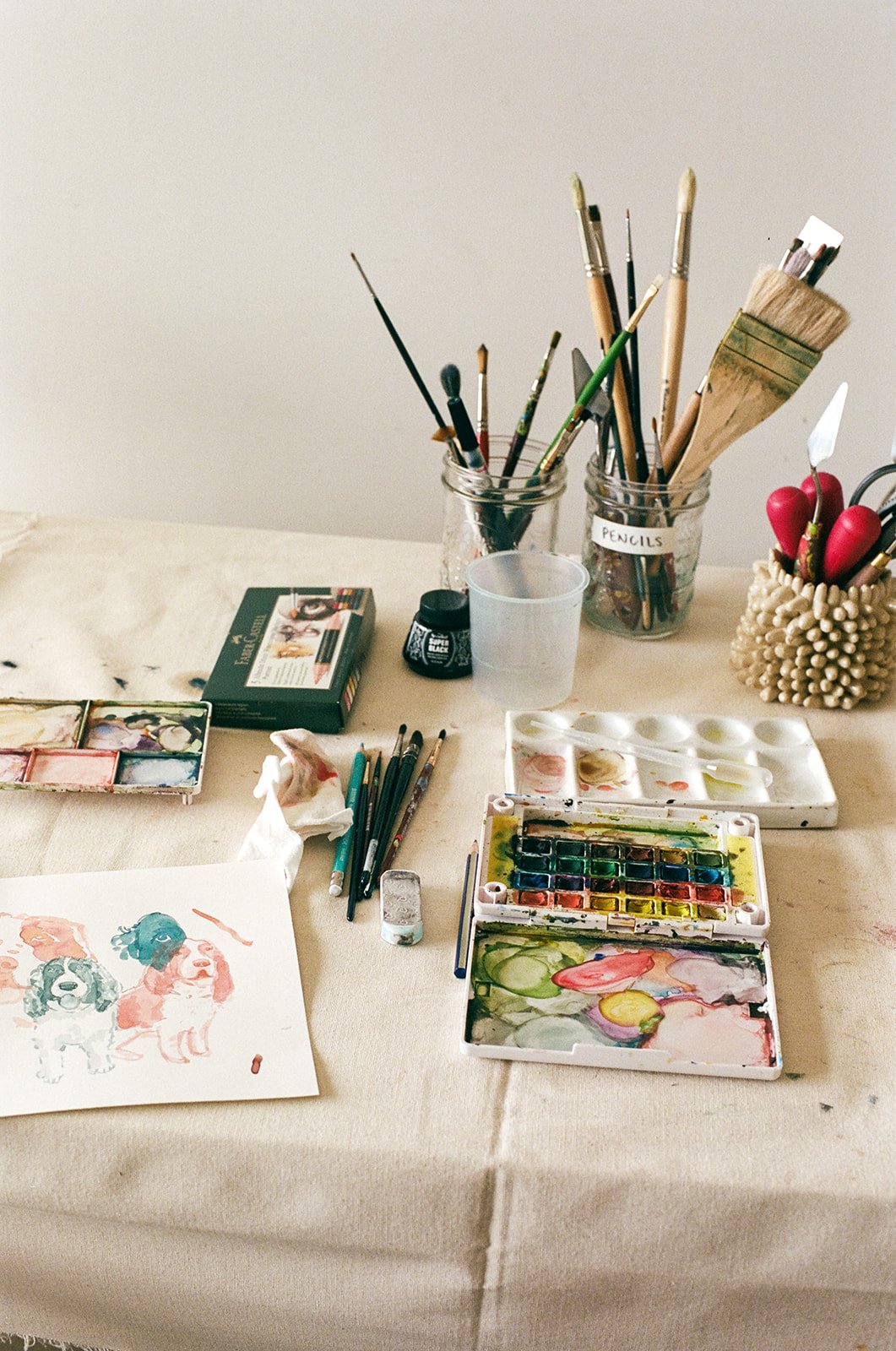 An art and painting supplies workspace on a beige tablecloth with watercolor paints, brushes, a watercolor paper with a dog painting, and various art tools.
