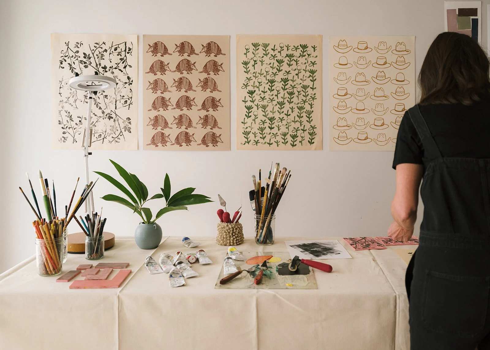 An artist's workspace with a table covered in art supplies including paint tubes, brushes, and other tools, and decorative posters with nature-inspired and animal patterns on the wall behind.