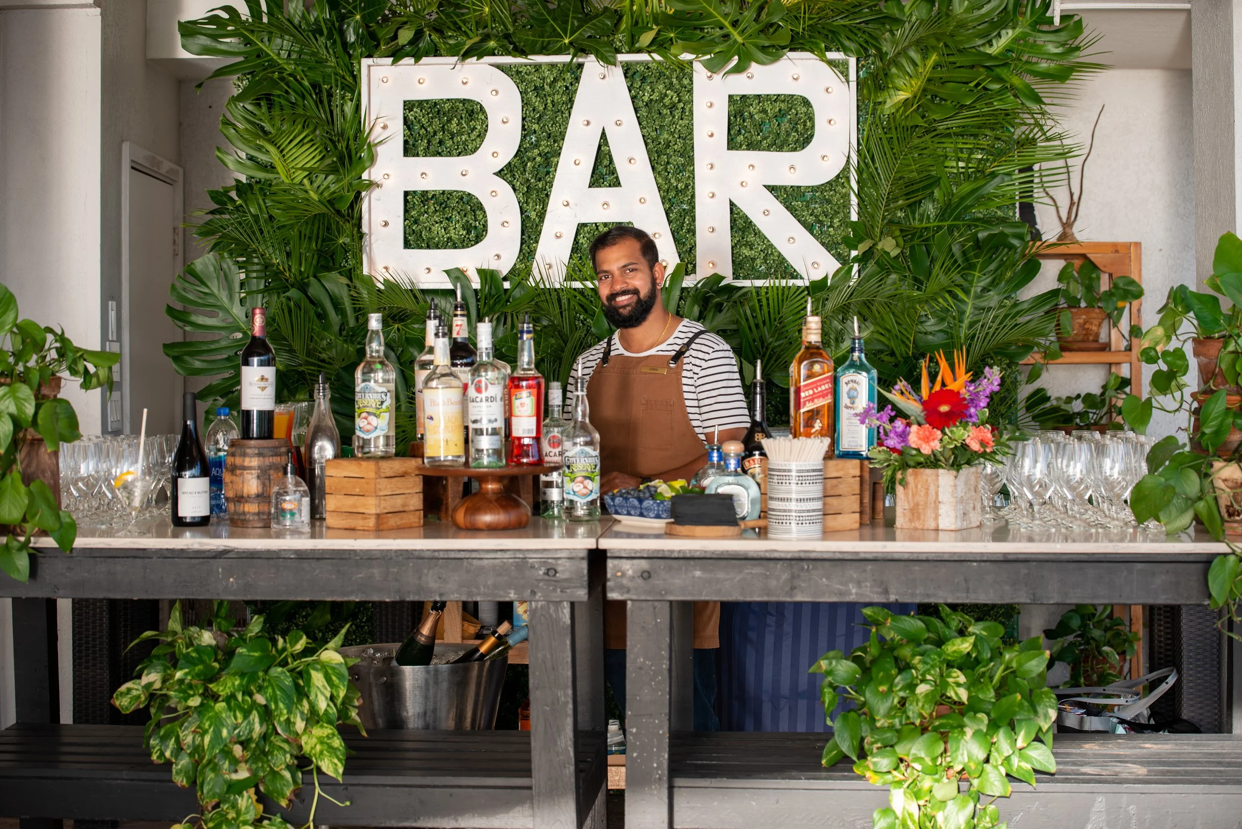 A bartender standing behind a bar counter with various bottles of alcohol, barware, and a flower arrangement. Large illuminated sign spelling 'BAR' is in the background, surrounded by green foliage and plants.