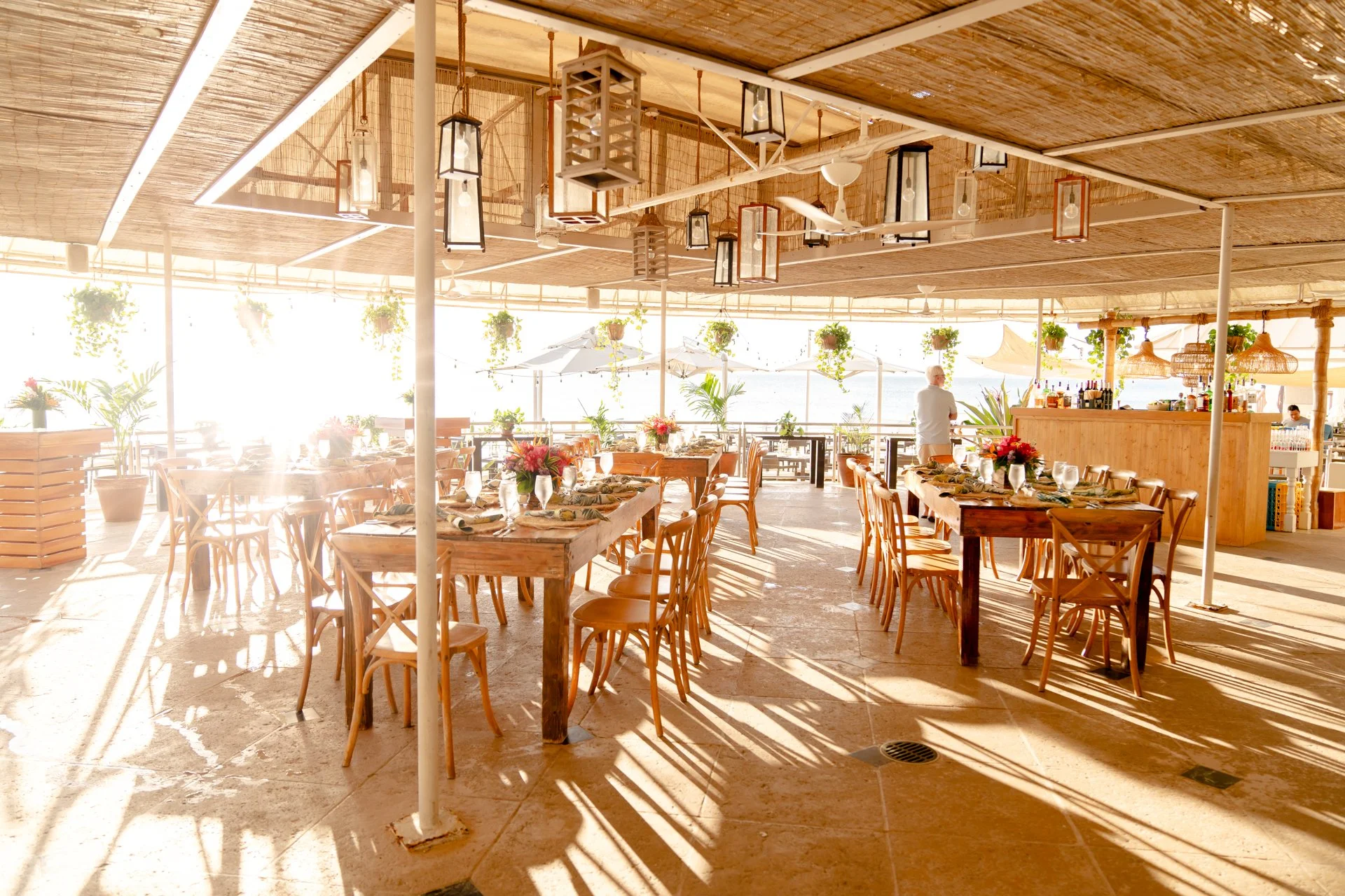 An open-air restaurant or event space with wooden tables and chairs, decorated with flowers. Hanging lanterns and plants are suspended from the ceiling. Sunlight streams in, creating bright reflections and shadows on the floor, with some umbrellas an