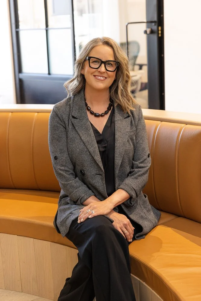 A smiling woman with shoulder-length blonde hair, glasses, wearing a navy blue jacket and layered necklaces.