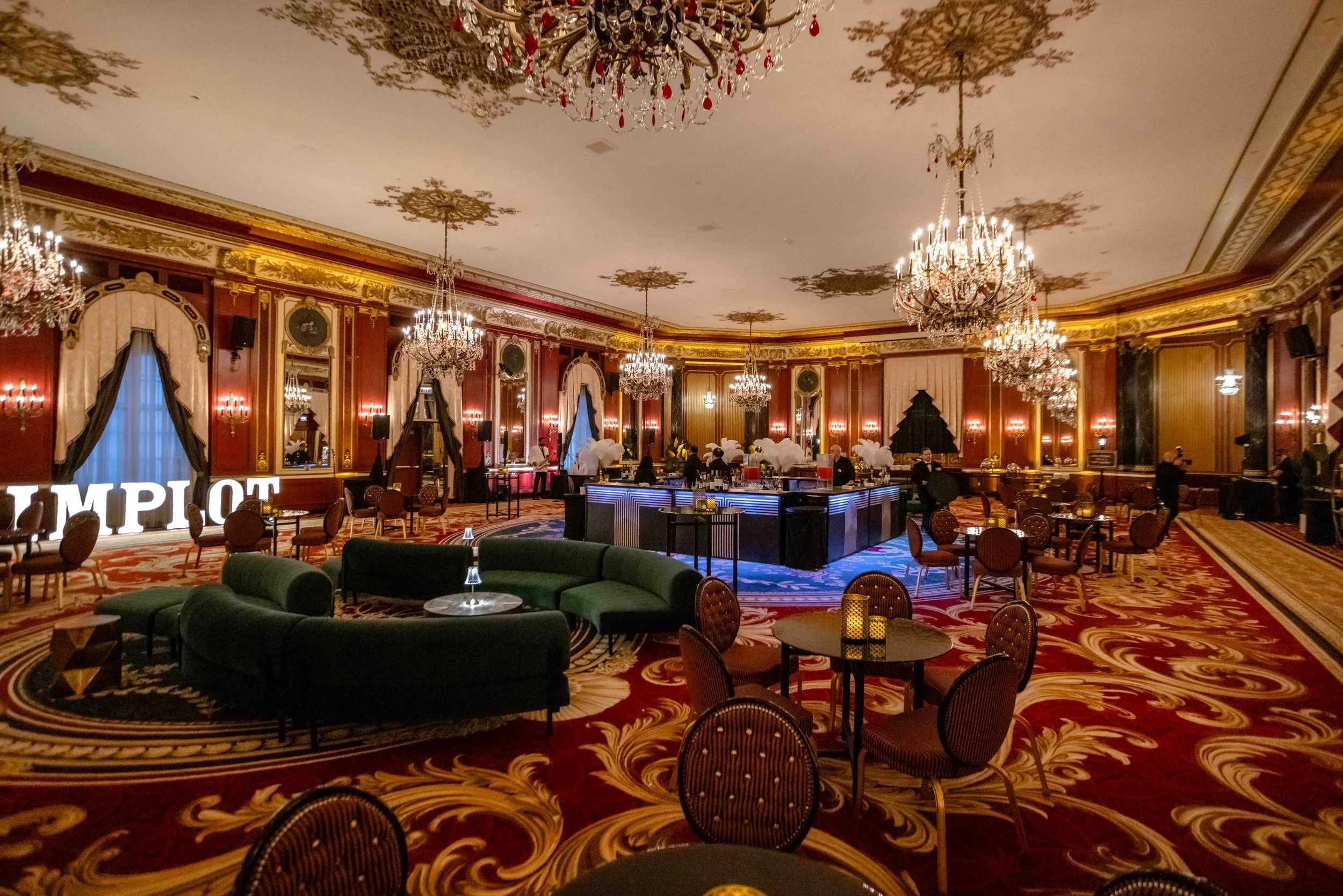 Elegant ballroom with chandeliers, red and gold decor, round tables with chairs, a green curved sofa, and a bar in the center.