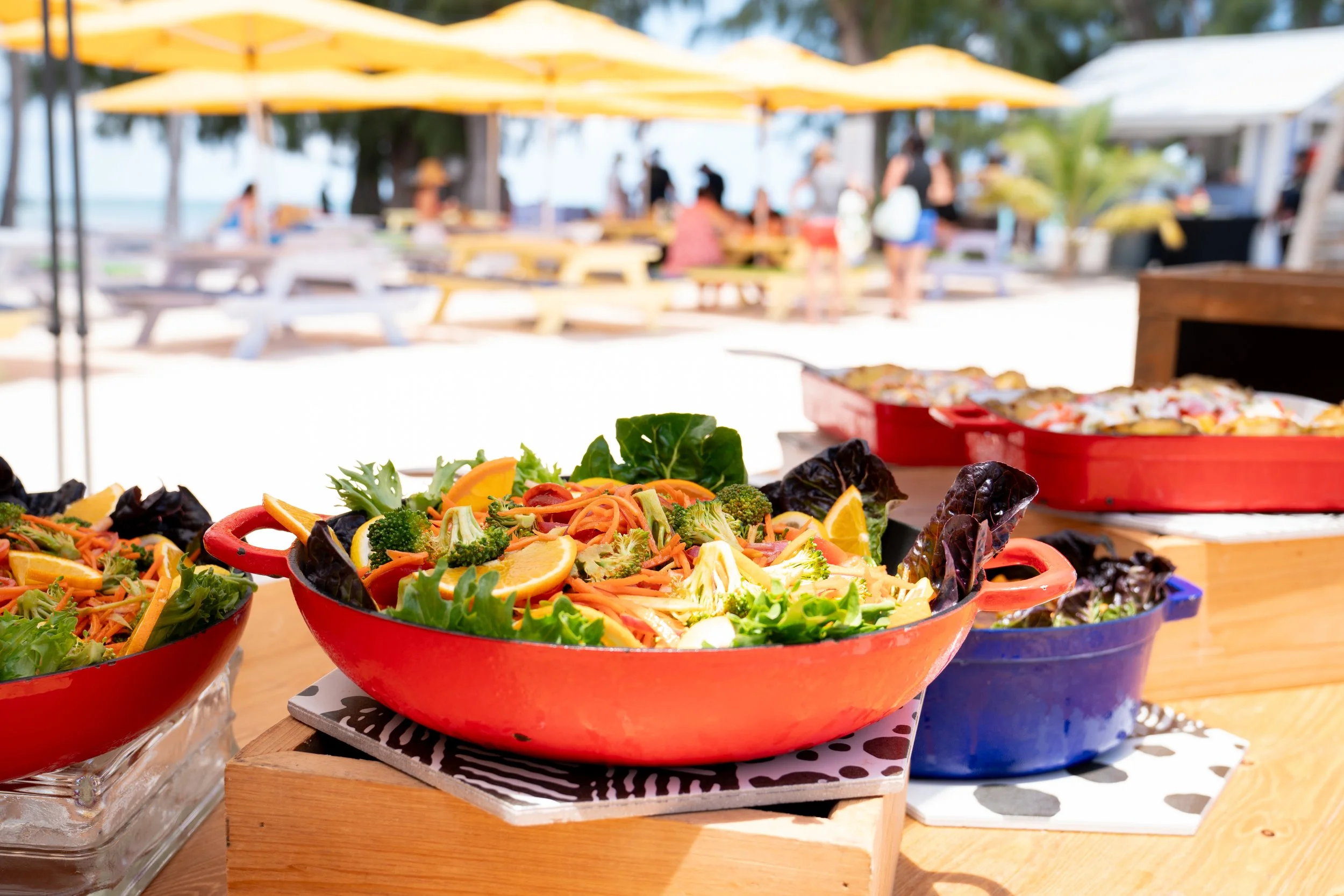 Colorful fresh salads with lettuce, carrots, broccoli, and orange slices in red and blue bowls at outdoor beachside gathering.