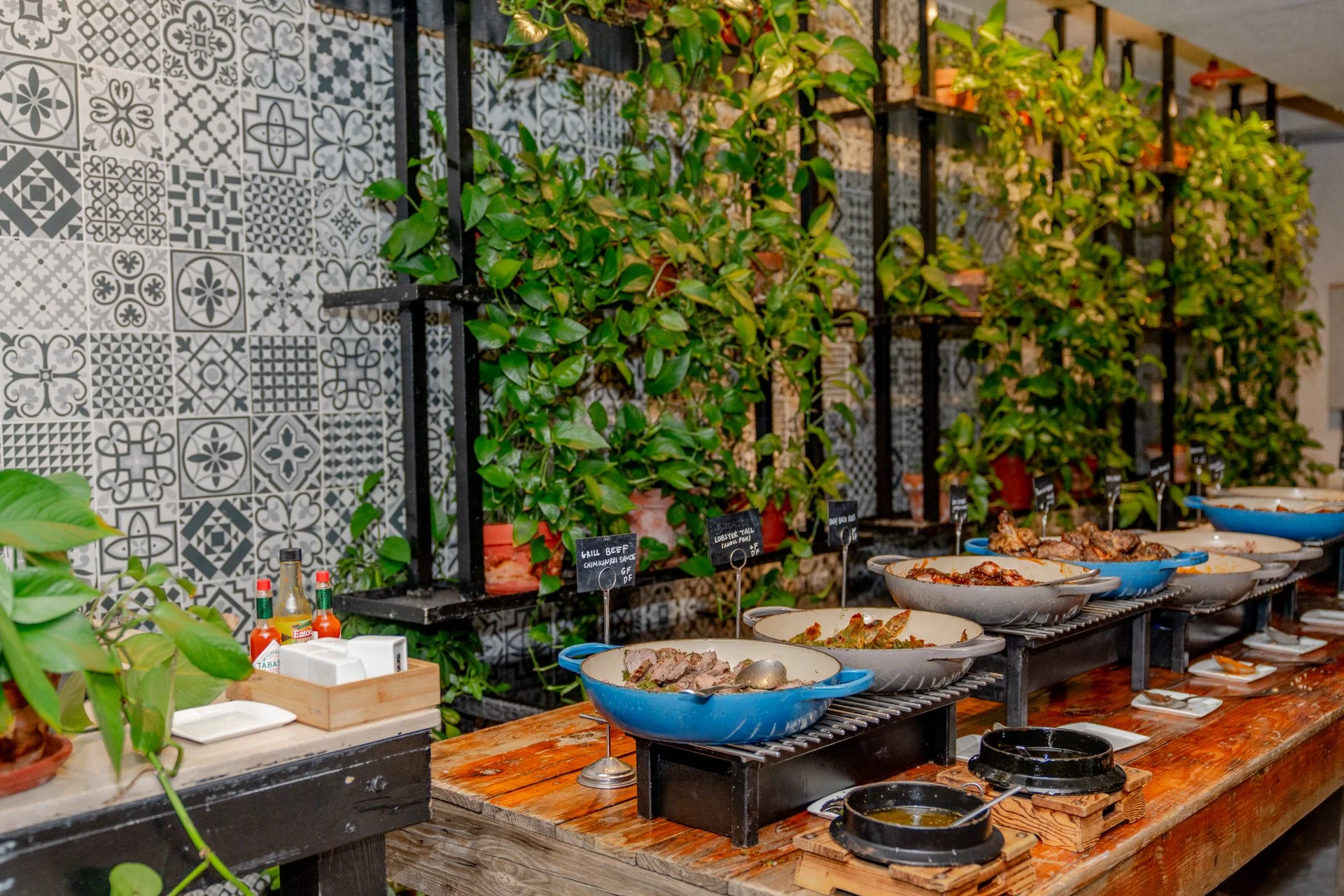 Buffet setup with food dishes in bowls on a wooden table, condiments, and potted plants against a decorative tiled wall.