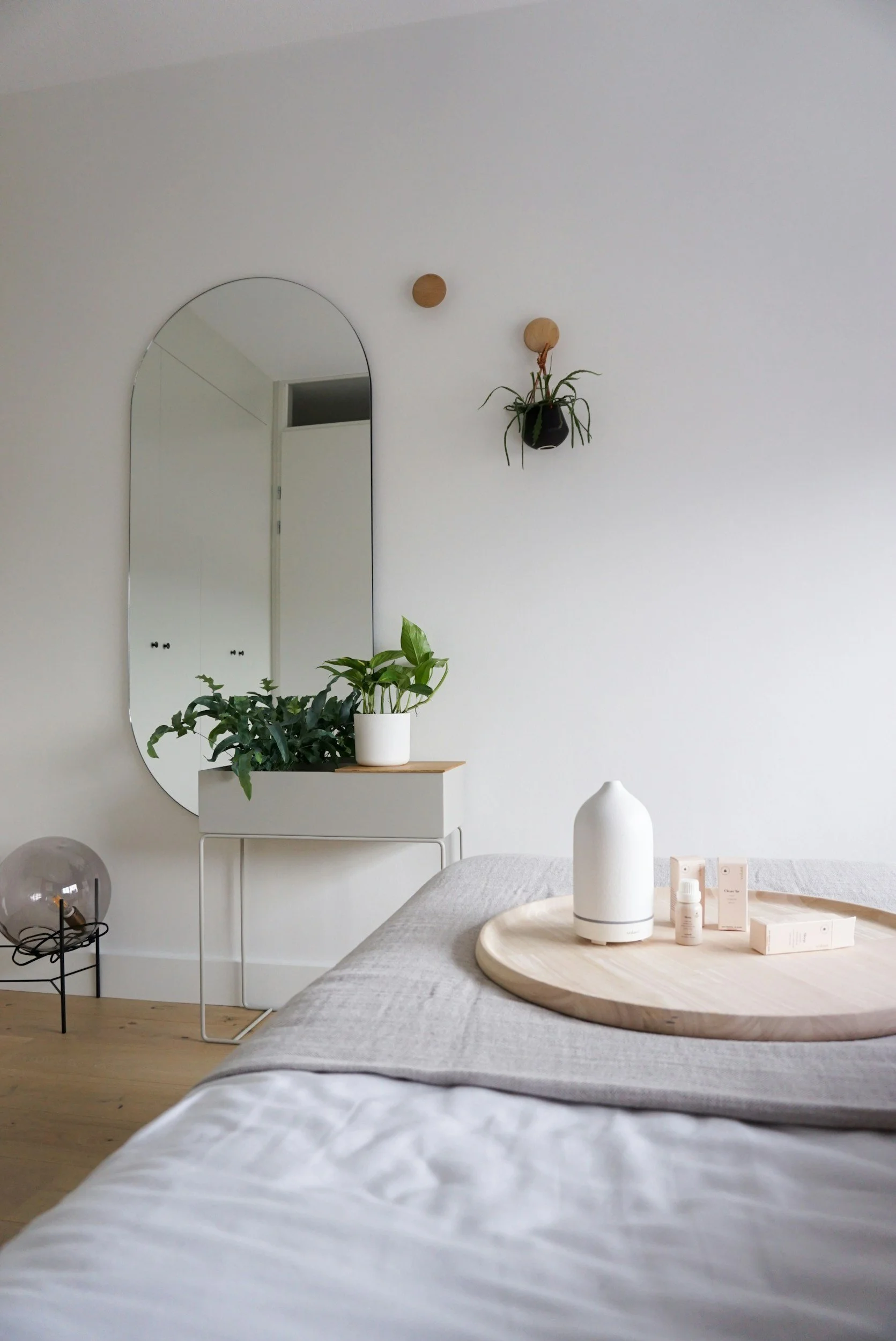 A minimalist bedroom with a white wall, an oval mirror, a small white table with potted plants, a bed with light bedding, a wooden tray with a diffuser, skincare products, and a small side table with a glass dome lamp.