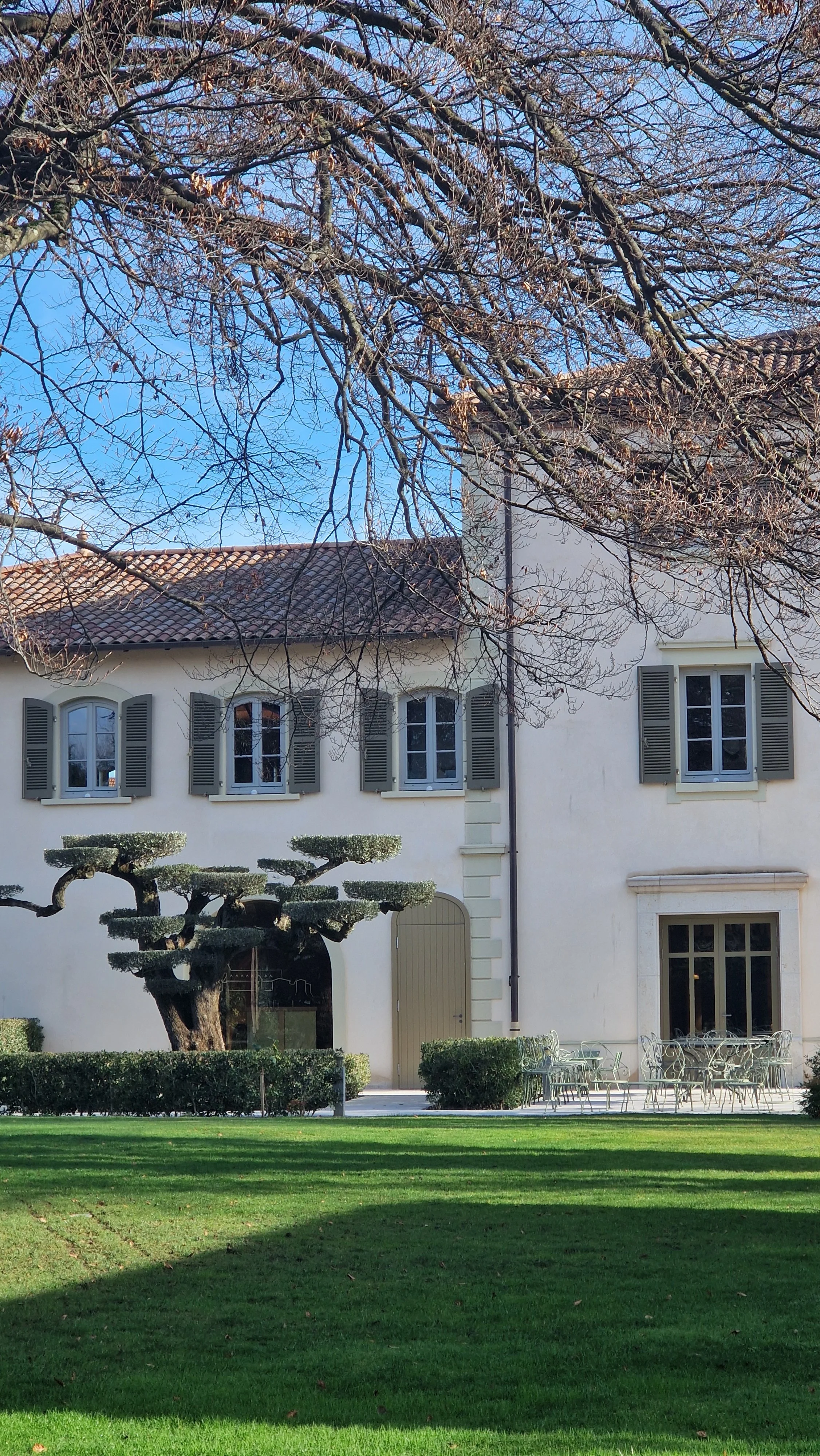 A white two-story house with gray shutters and a red-tiled roof, surrounded by a well-manicured lawn and trimmed bushes, with tall leafless trees and a clear blue sky overhead.
