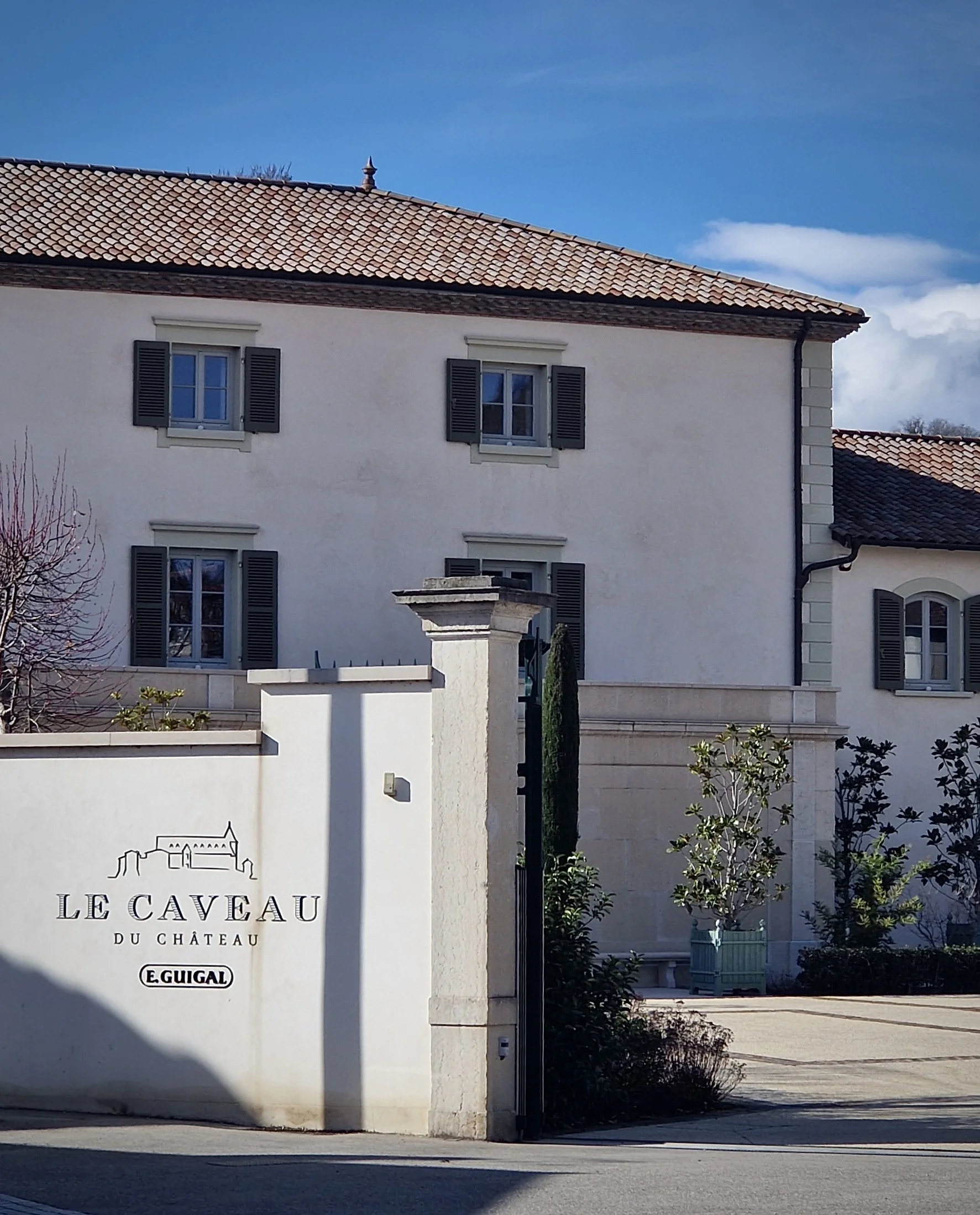Exterior of a white building with a tiled roof, windows with black shutters, and a sign that reads 'LE CAVEAU DU CHATEAU E. GUIGAL' with a simple outline of a castle.