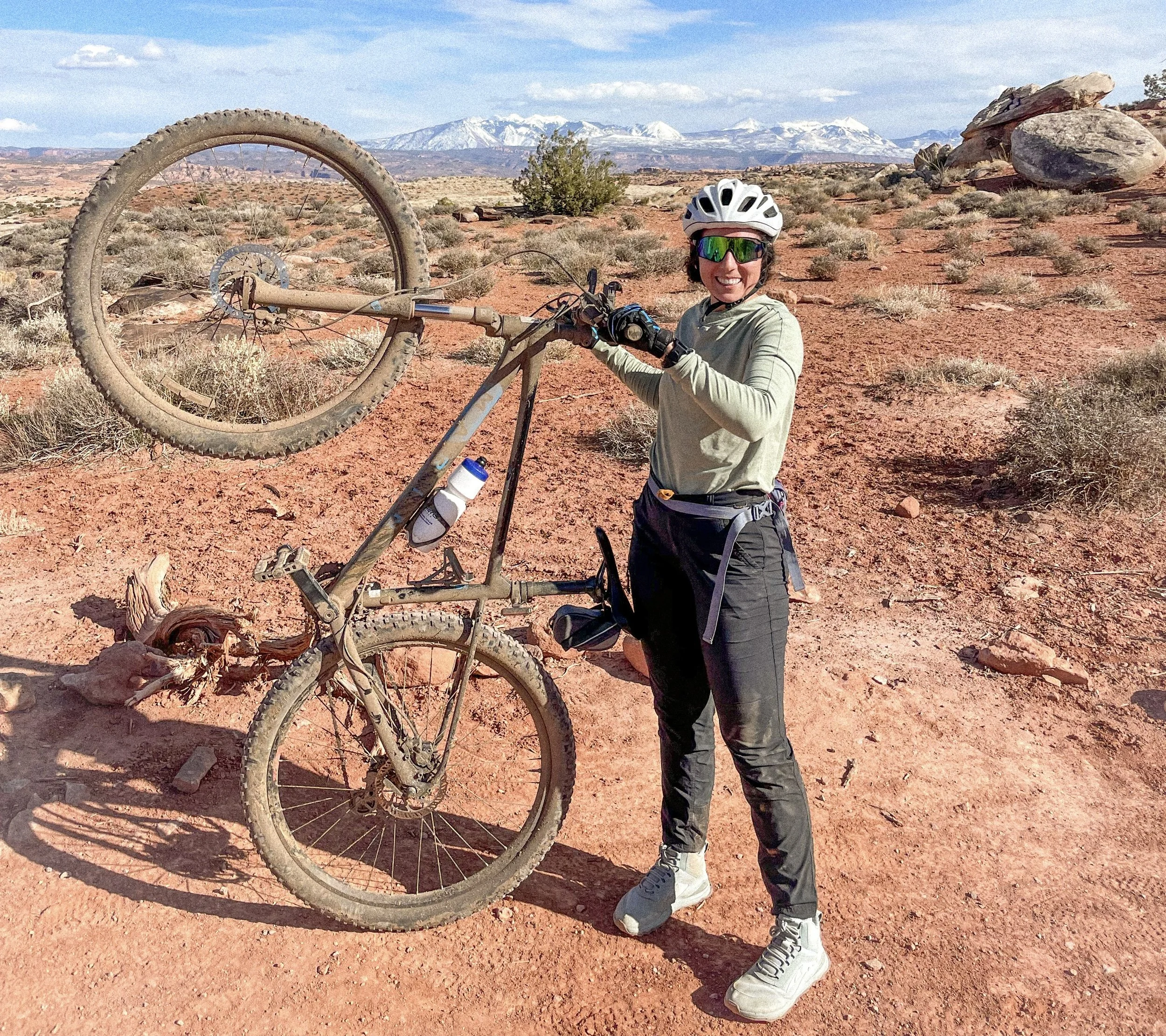 A woman in outdoor athletic clothing holding a mountain bike in a desert landscape with red sand, rocks, and shrubs, mountain range with snow-capped peaks in the background, and a partly cloudy sky.