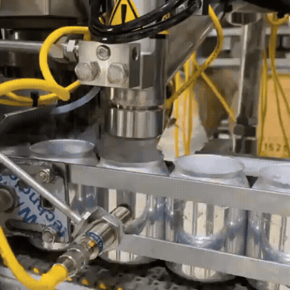 Primer plano de una línea industrial de enlatado: un cabezal metálico (tipo llenado/sellado) trabajando sobre latas alineadas en una banda transportadora, con mangueras amarillas y estructura de acero inoxidable en un entorno de planta.