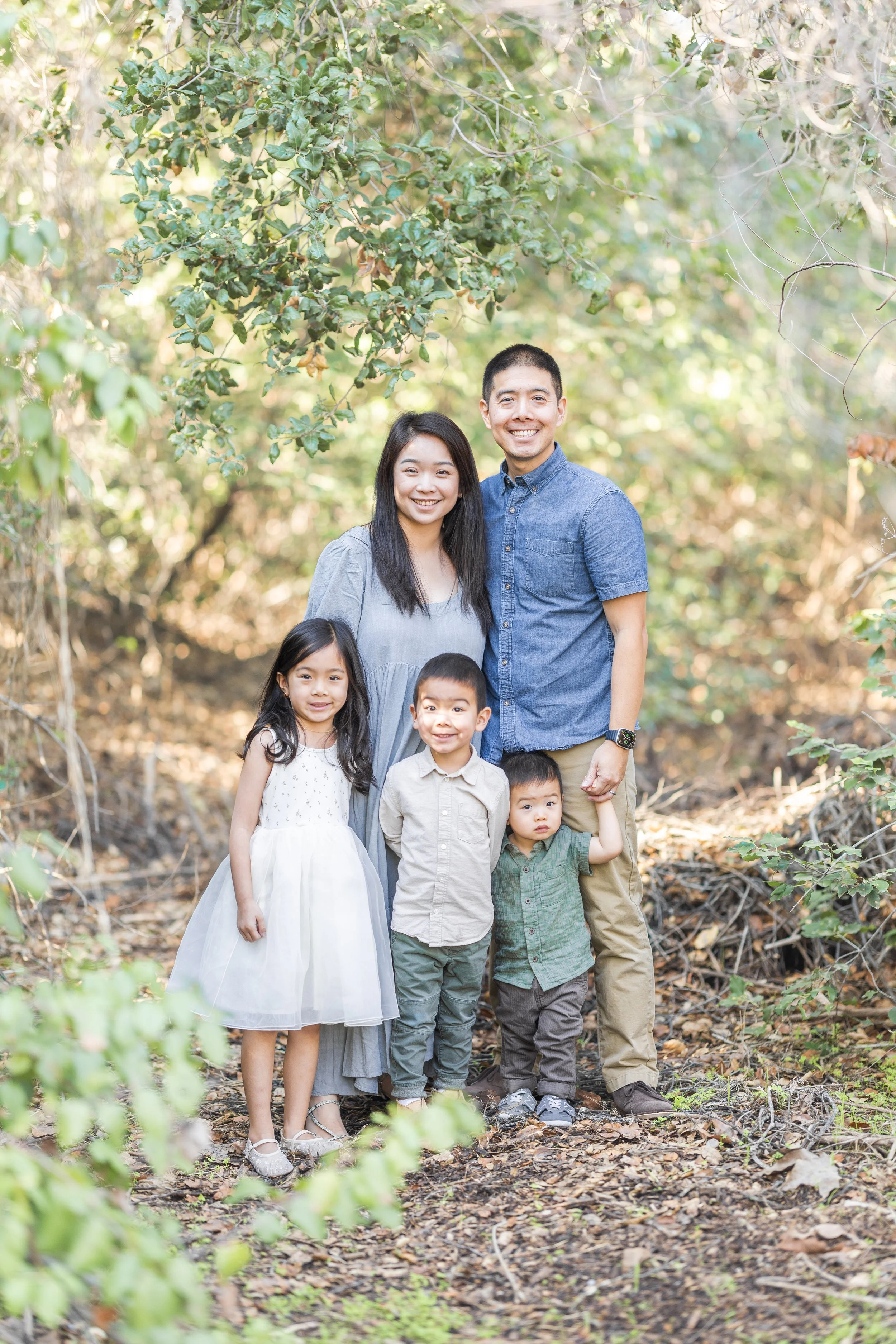 A family of five outdoors, standing on a forest trail surrounded by green foliage, smiling at the camera.